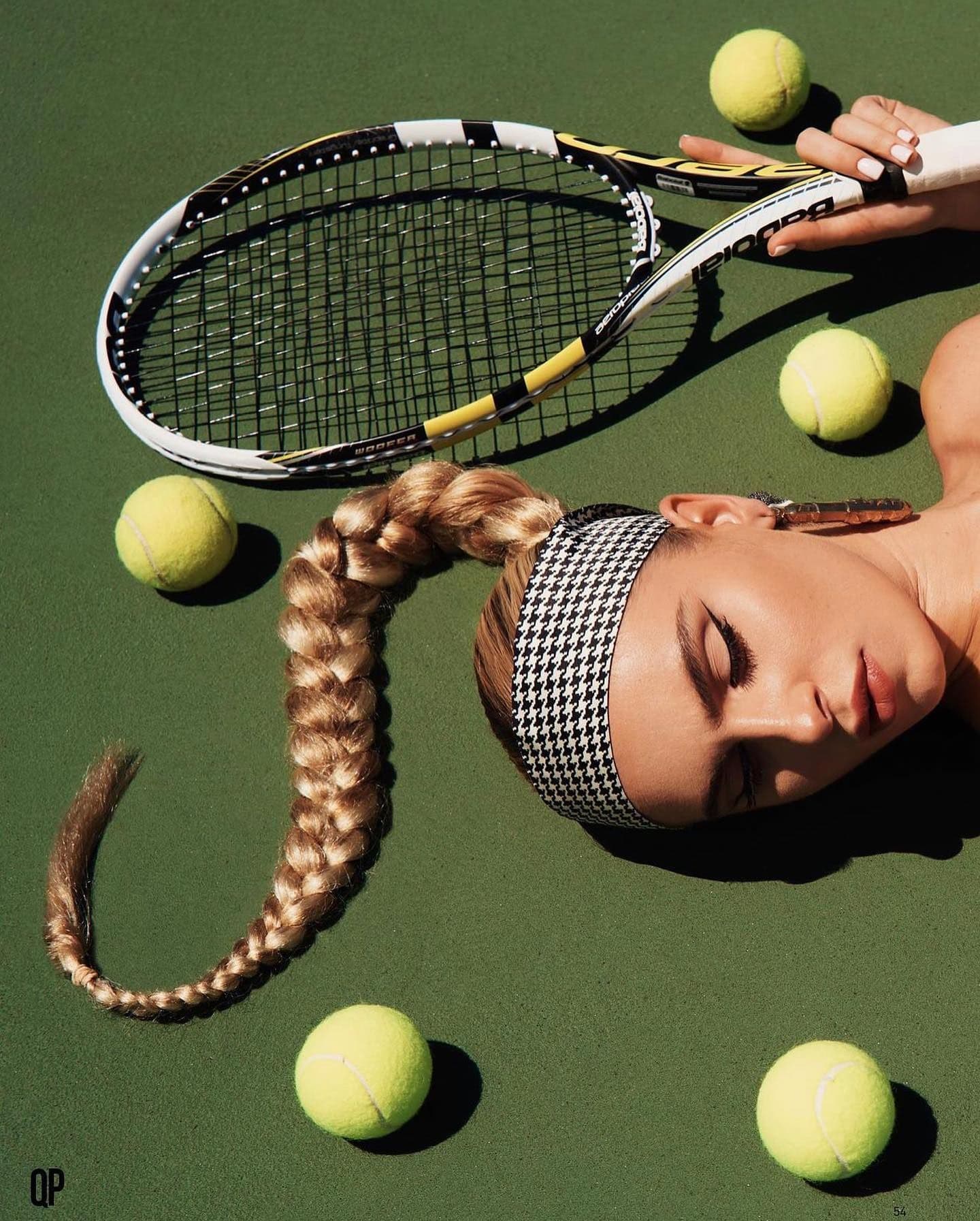 Editorial photo featuring a blonde model with long braid, tennis racket, five tennis balls lying on a green tennis court.