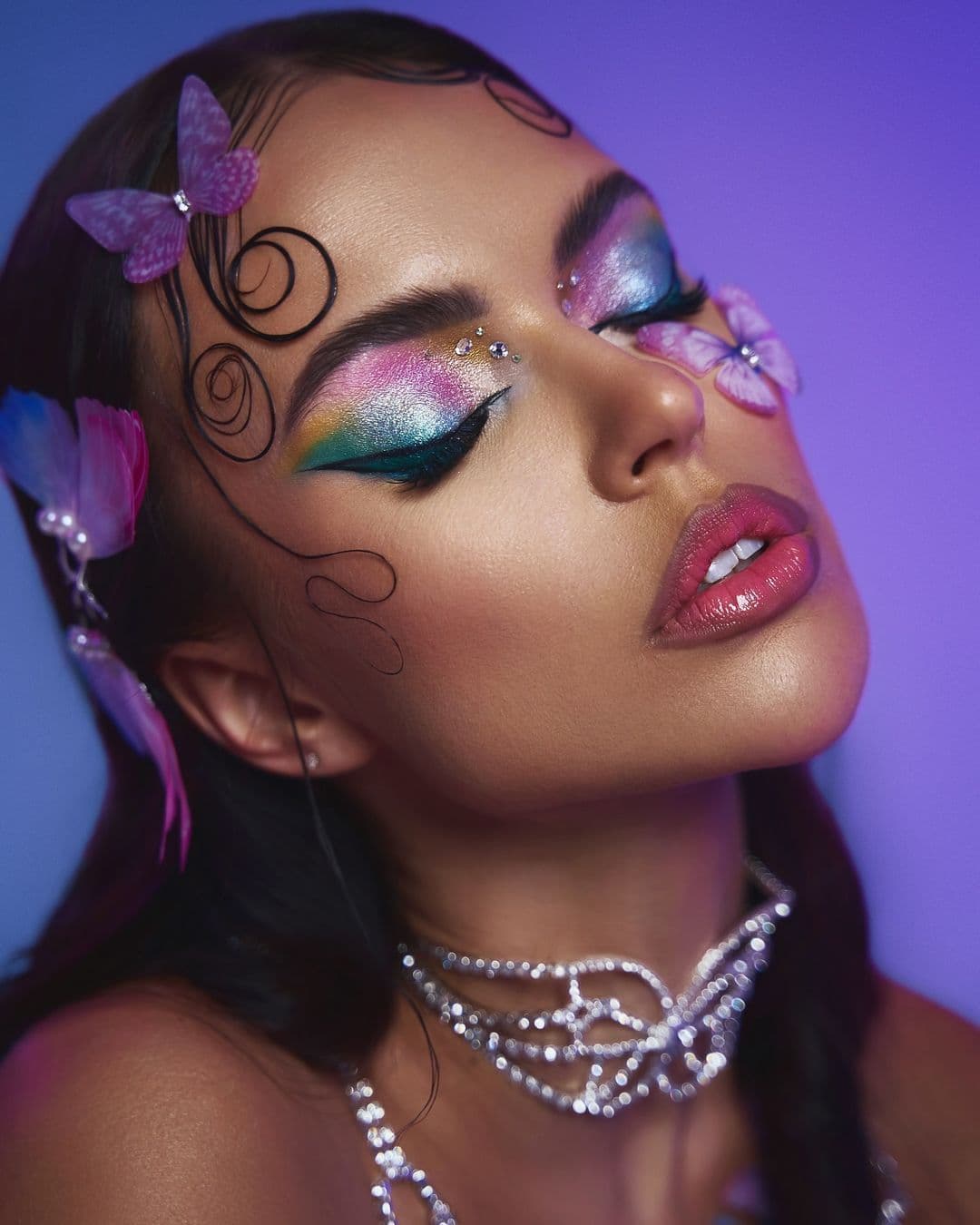 Woman with artistic makeup, intricate curly hair, butterflies, and necklace poses with eyes closed.