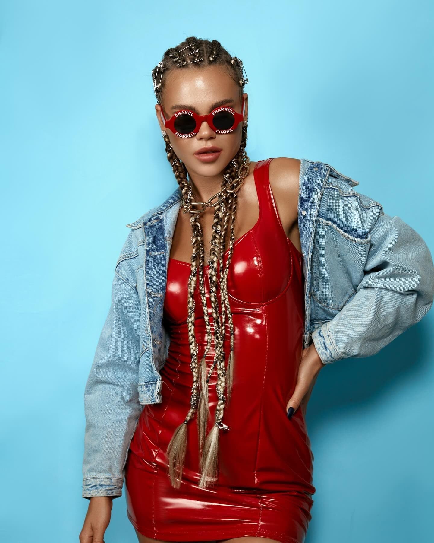 A woman in braids, a red dress, and red designer sunglasses poses in a denim jacket in a studio with a light blue backdrop.