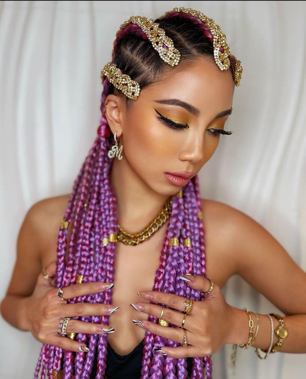 A woman with purple braided hair extensions styled in cornrows with gold embellishments, makeup, jewelry and manicured nails.