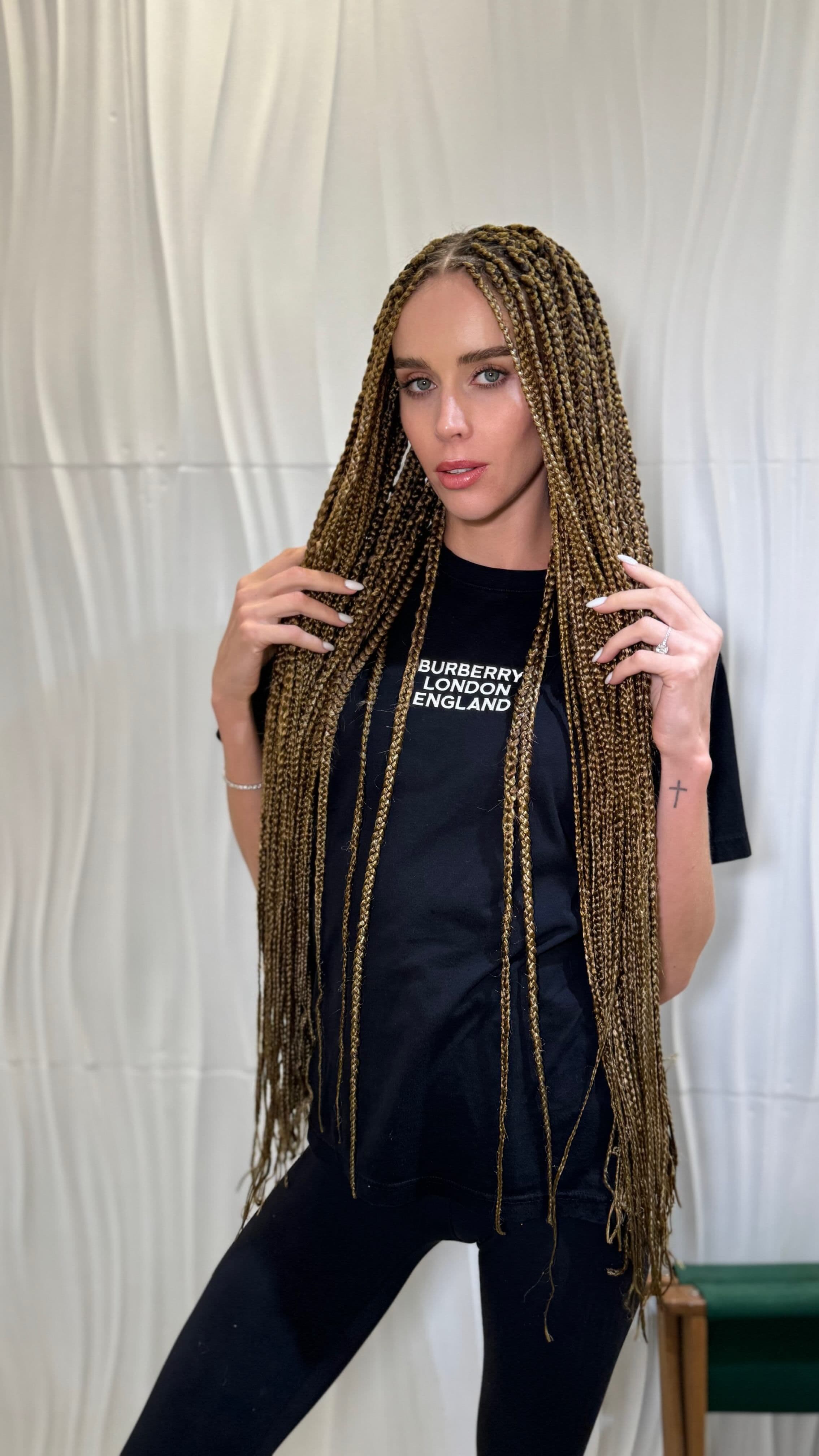 Woman with long, light brown box braids wearing a black shirt and leggings posing in front of a textured wall.