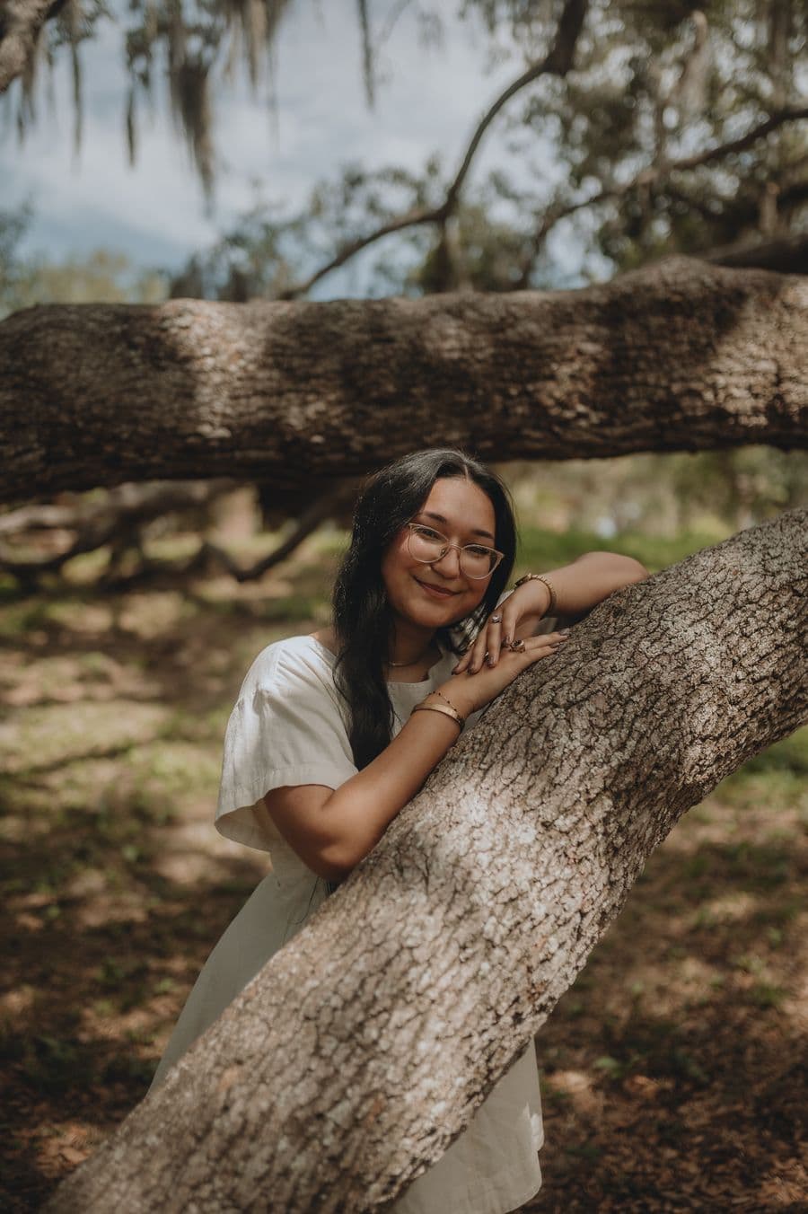 A creative portfolio portrait of a woman with long dark hair and glasses leaning against a large oak tree branch in a sun-dappled park.