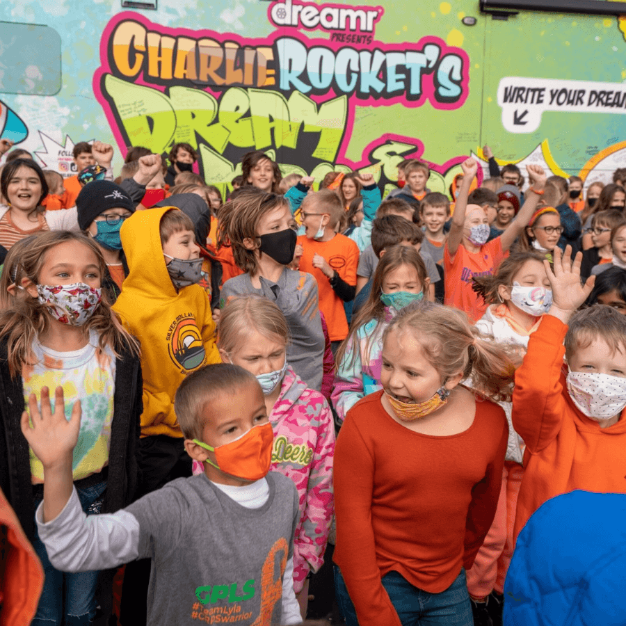 A large group of elementary-aged children in masks gather in front of a 'Charlie Rocket's DREAM' bus.