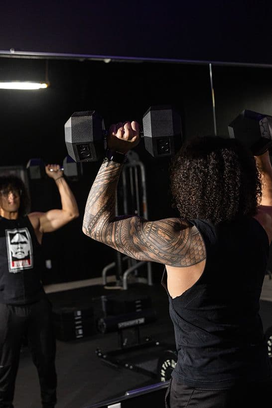Trainer Joel lifts dumbbells overhead, reflected in a mirror. He specializes in stretch therapy and mobility.