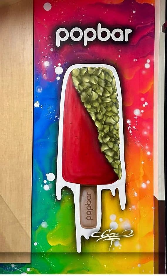 A commercial street art mural for Popbar, featuring a large red popsicle with a green pistachio topping on a vibrant rainbow wall.