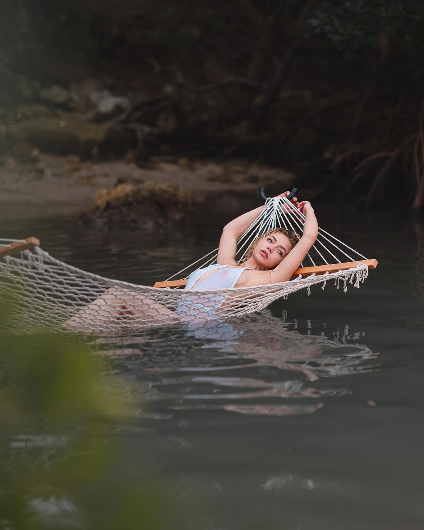 A model poses in a white hammock suspended over water, arms raised, for a glamour photoshoot.
