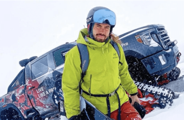 Adventure influencer Tristan Hamm stands in the snow with his snowboard, next to a truck modified with snow tracks.