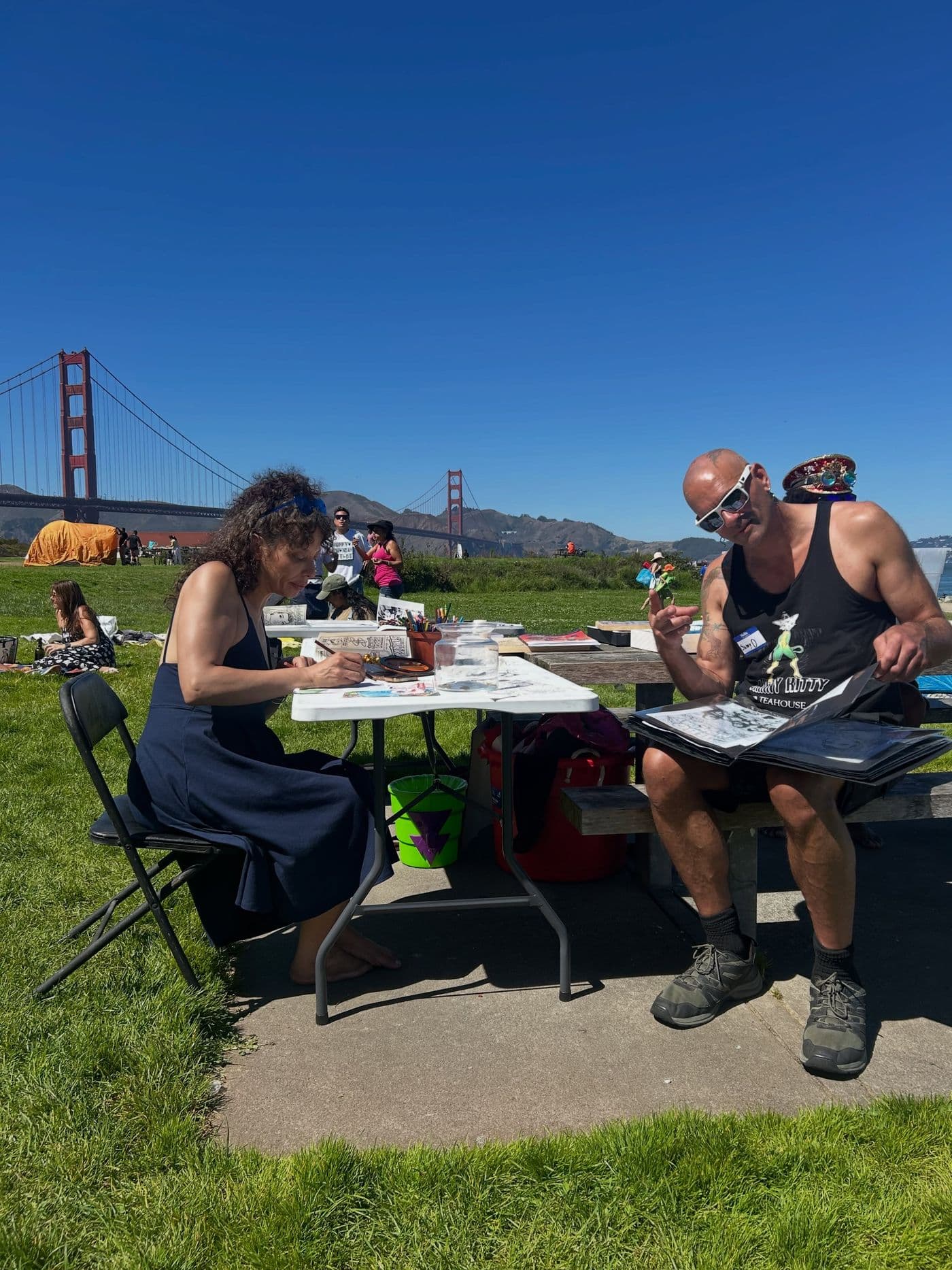 A creative portfolio session outdoors where a woman draws at a white table and a man reviews a physical visual portfolio with the Golden Gate Bridge in the background.