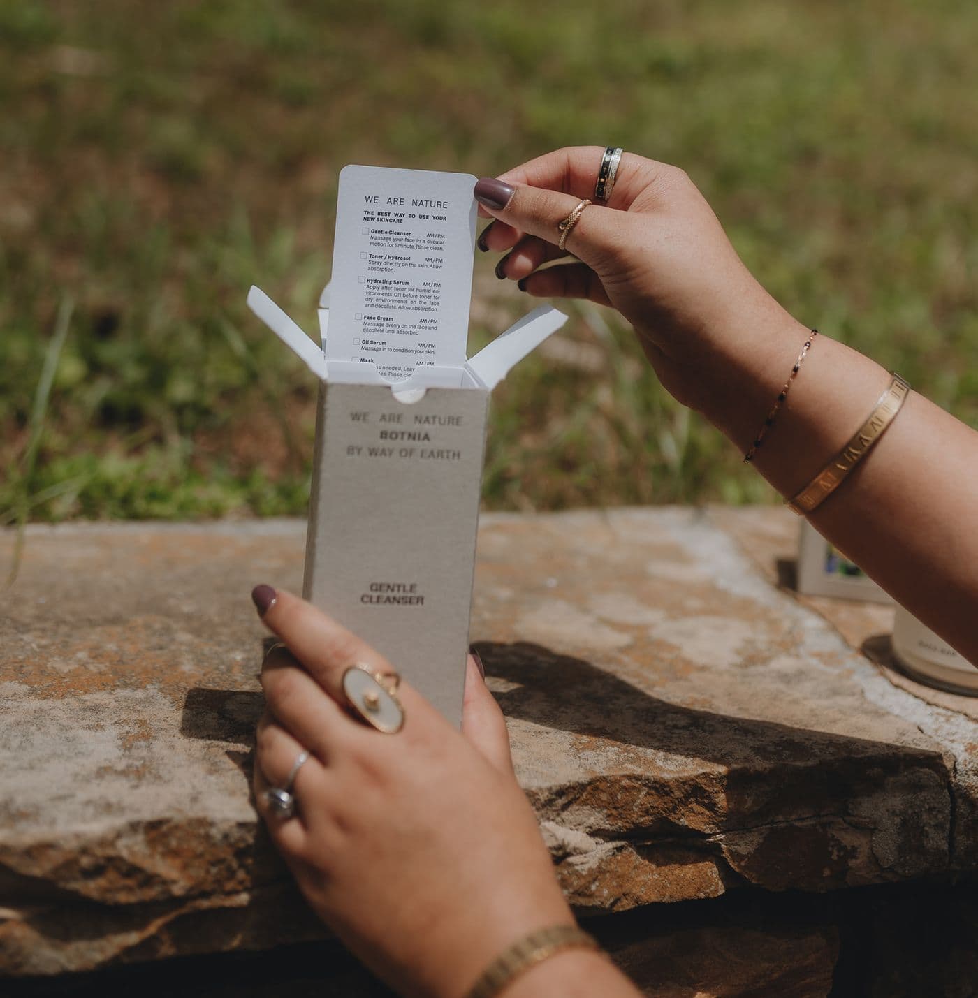 A person with gold jewelry pulls a skincare instruction card out of a Botnia Gentle Cleanser box while sitting outdoors on a stone ledge.
