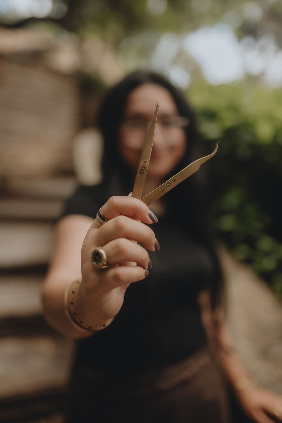 A lash artist holds up two gold professional tweezers engraved with names, showcasing her tools for a beauty portfolio.