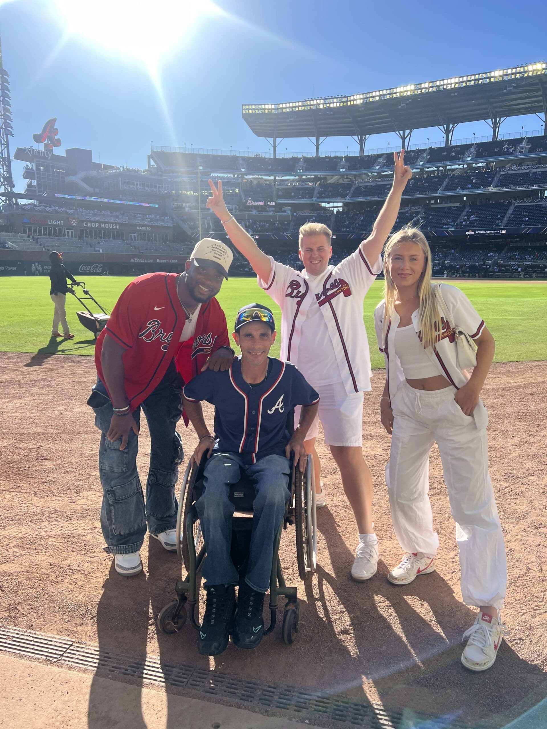 Charlie Rocket poses with fans in Braves jerseys on a baseball field, one in a wheelchair, on a sunny day.