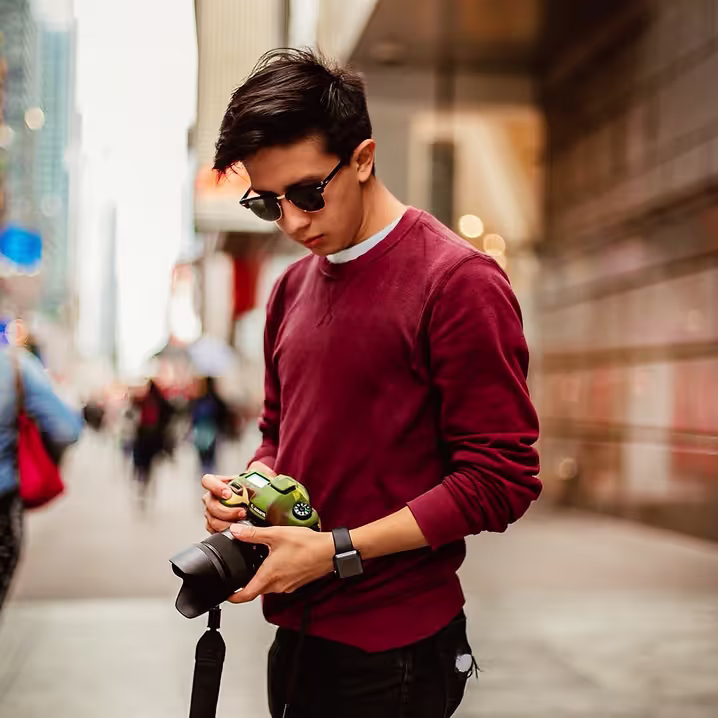 Erick FernandoQ holding a DSLR camera and wearing a maroon long sleeve shirt, sunglasses and black pants.
