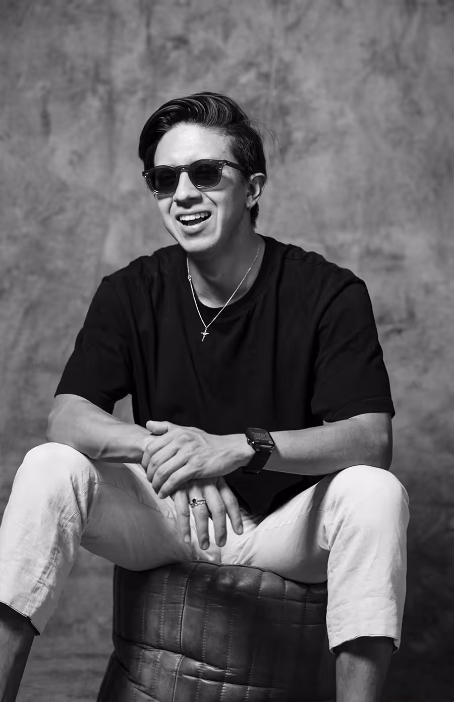 Black and white photo of Erick Fernandoq in sunglasses sitting with hands clasped and smiling.