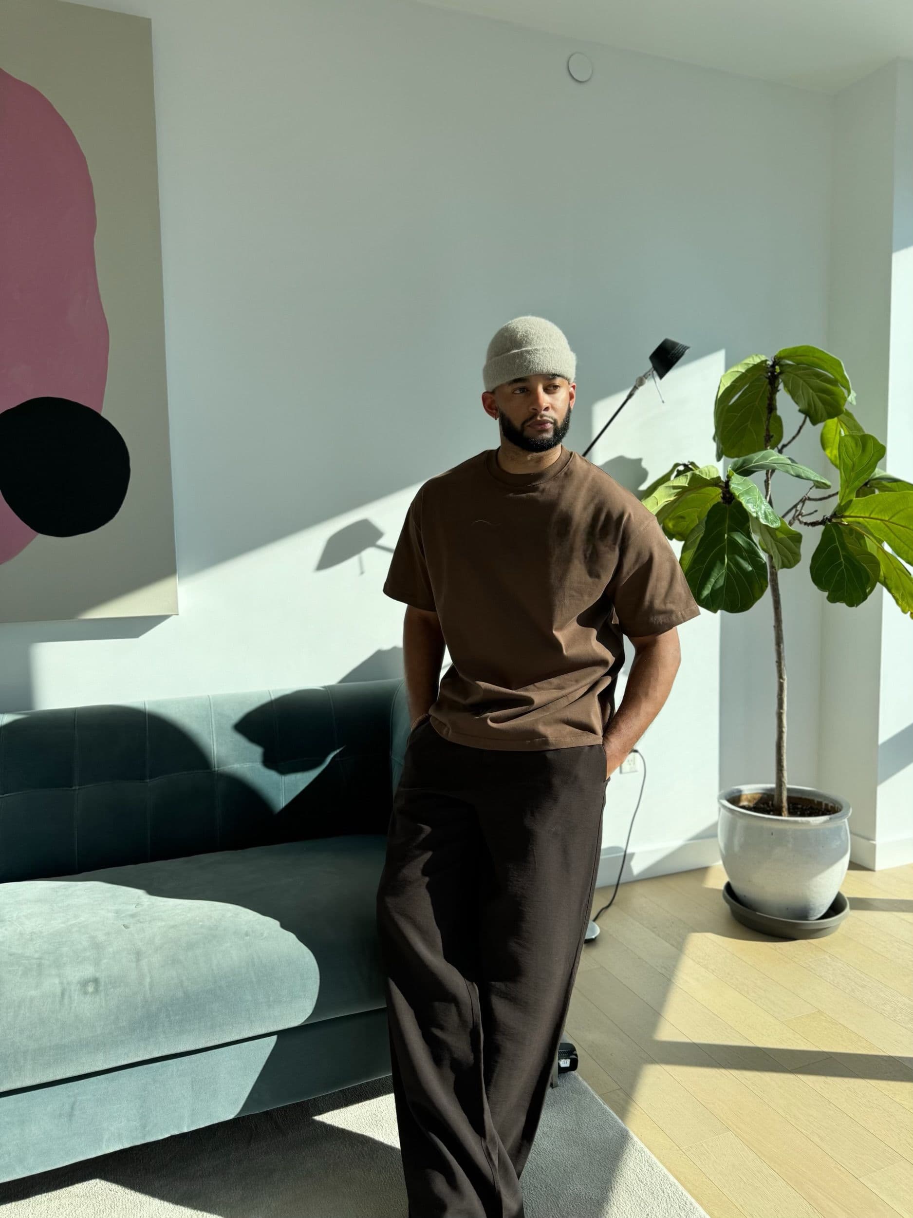 Creative Director Mike Press, wearing a brown t-shirt and beanie, leans against a teal sofa in a sunlit modern room.