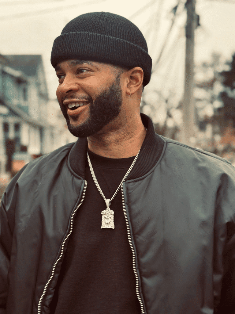 Mike Press, a man with a beard, wears a black beanie and bomber jacket, smiling and looking left.