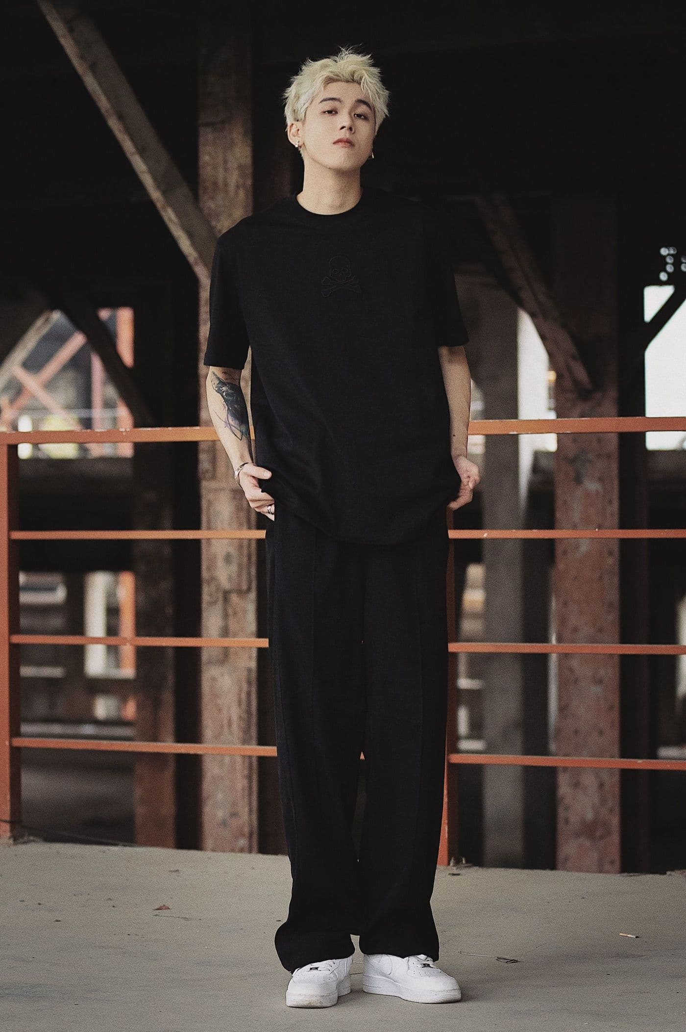 Fashion-forward man with dyed hair and tattoos posing outdoors in black attire.