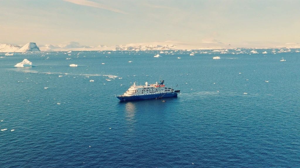 A cruise ship navigating through icy Antarctic waters with scattered icebergs, against a backdrop of snowy mountains.