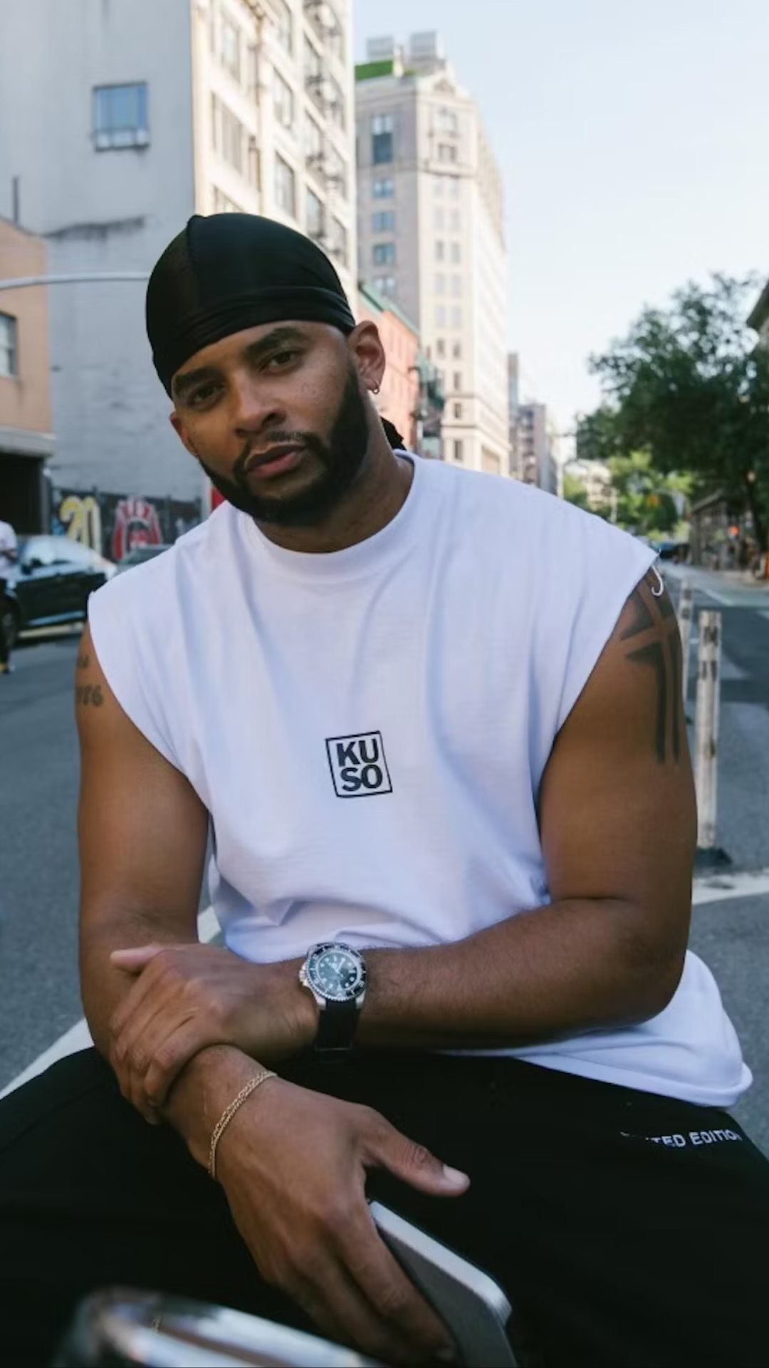 Bearded man with arm tattoos wearing a black skull cap and white sleeveless shirt with KUSO branding.