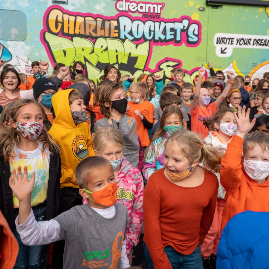 A large group of elementary-aged children in masks gather in front of a 'Charlie Rocket's DREAM' bus.