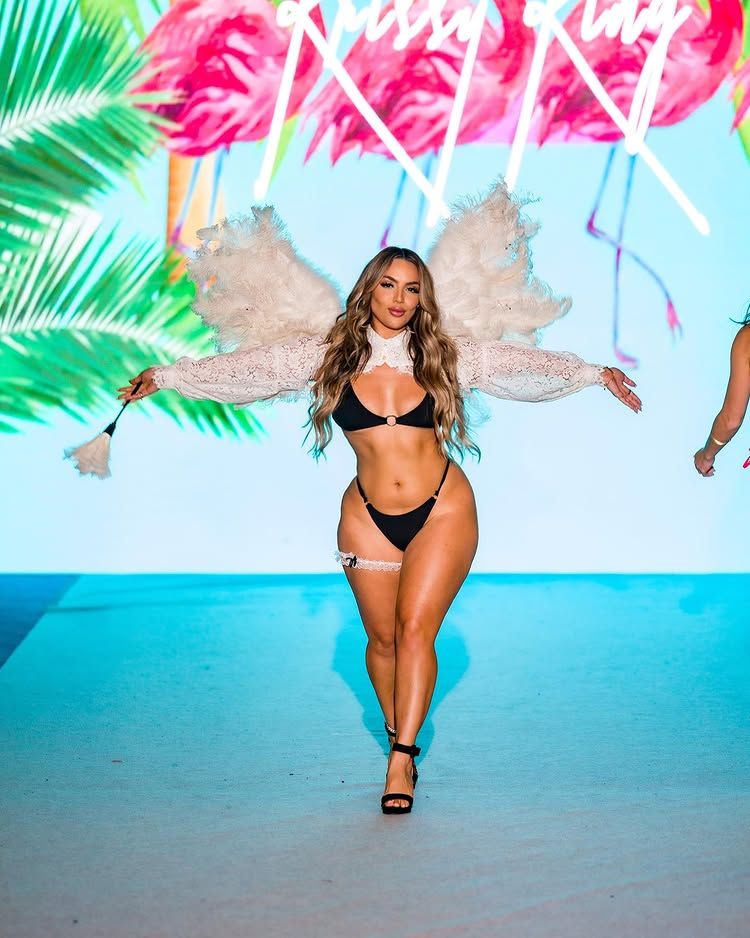Model Ara Bozorgian on a runway in black bikini, white lace winged top, and black heels, with flamingo graphics in the background.