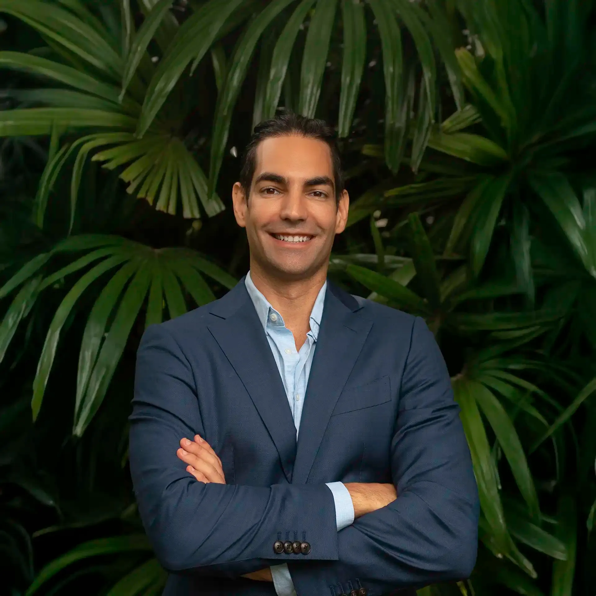 A photo of David Runyon smiling while crossing his arms in front of many tropical palm leaves.