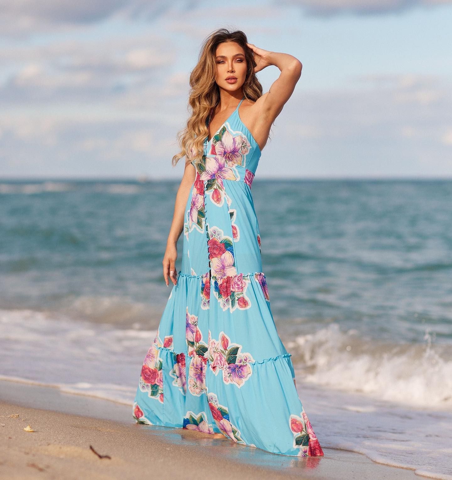 A model in a blue floral dress poses gracefully on a beach, with ocean waves around her feet.