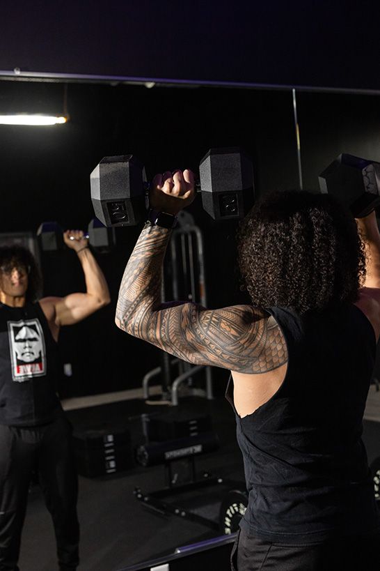 Trainer Joel lifts dumbbells overhead, reflected in a mirror. He specializes in stretch therapy and mobility.
