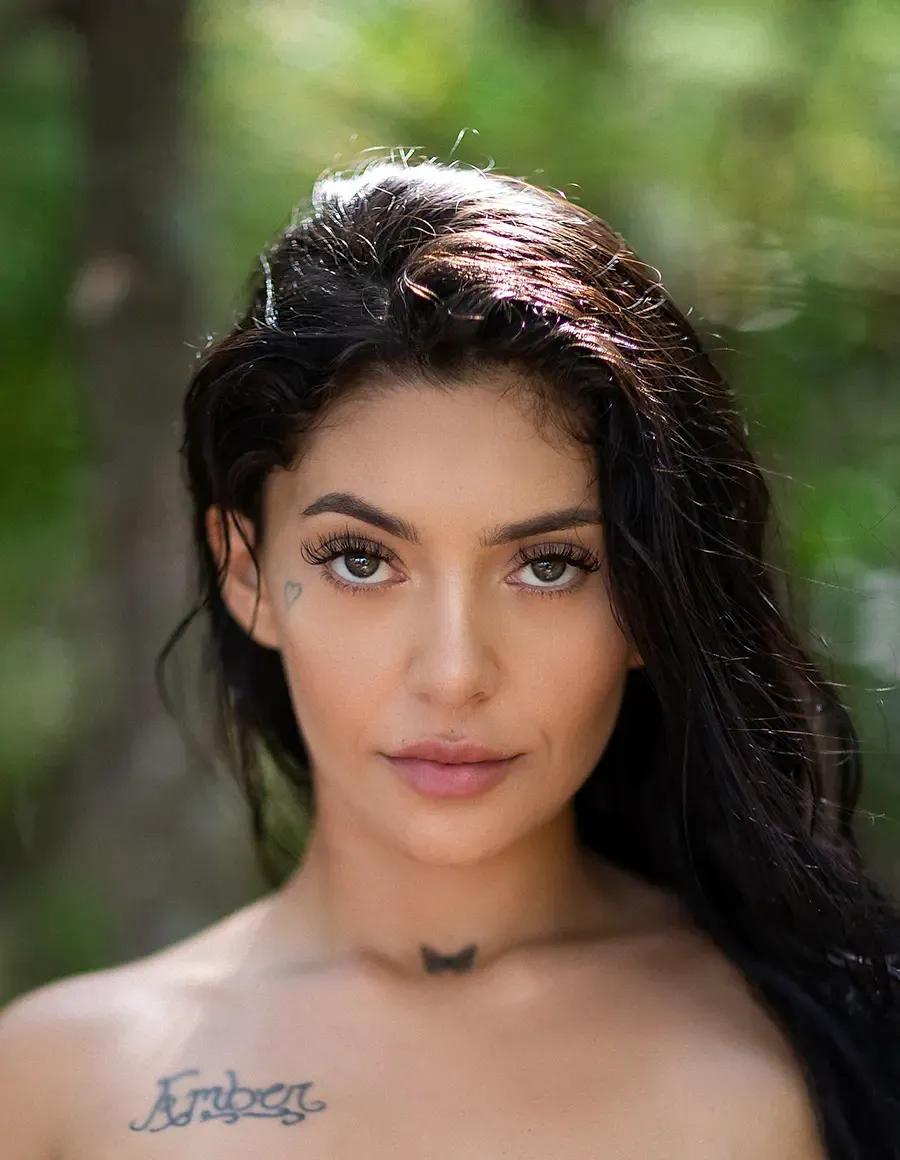 Model with dark hair and a heart tattoo near her eye, a butterfly on her throat, and 'Amber' on her chest.