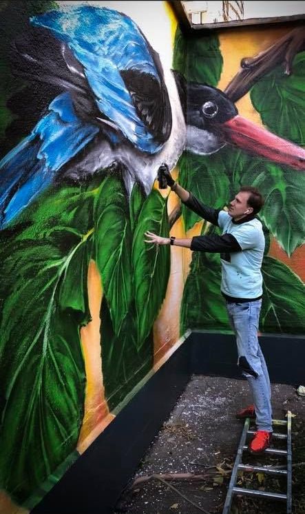 Muralist G.Z. Art stands on a ladder spray painting a large, colorful mural of a bird with blue wings on a client's wall.