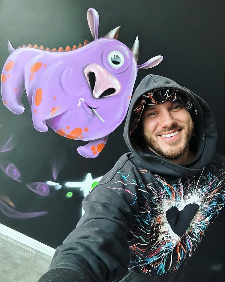 Oahu muralist from G.Z. Art smiles in front of his custom wall mural featuring a whimsical purple monster.