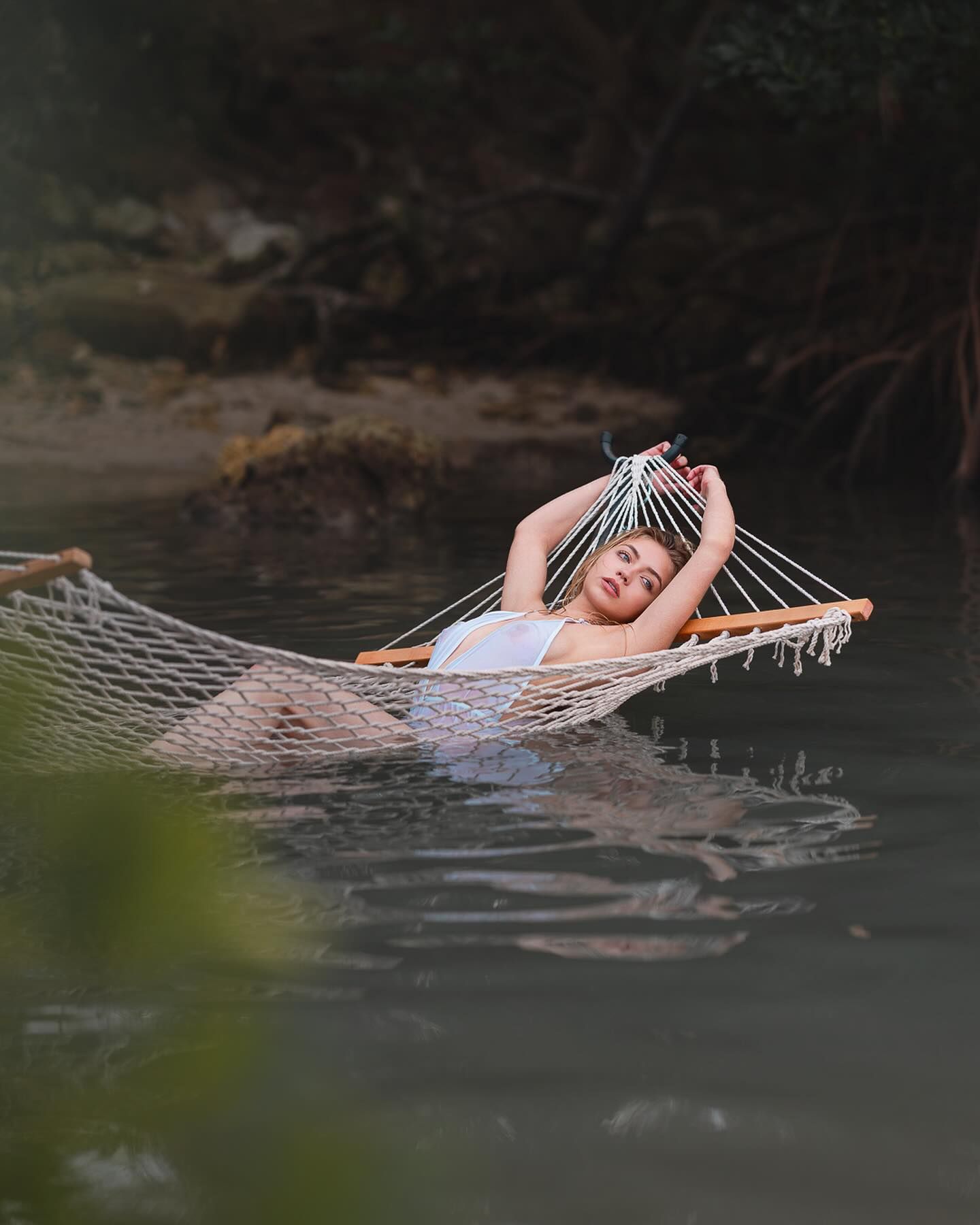 A model poses in a white hammock suspended over water, arms raised, for a glamour photoshoot.