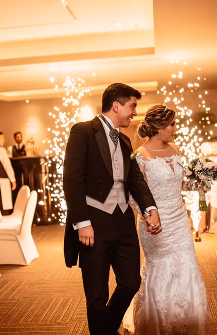 A newly married couple hold hands on the dance floor while fireworks sparkle behind them.